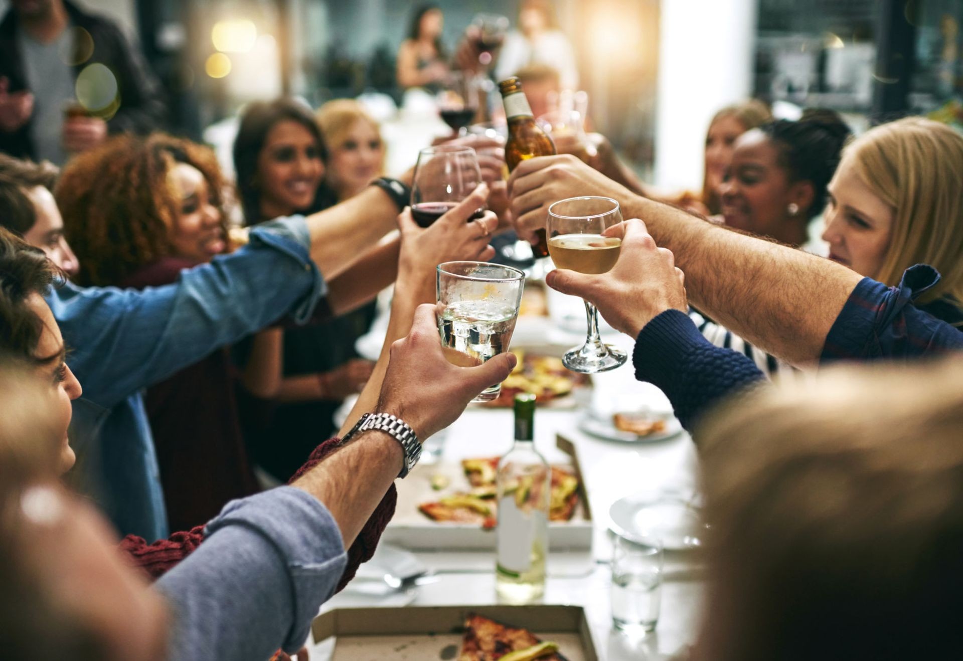 Group of people raising glasses in a toast at a dining table filled with pizza and wine bottles.